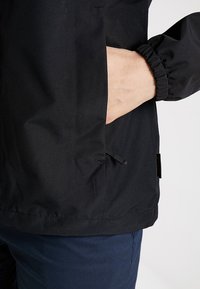 Svart vindjacka med dragkedja ficka, elastiska ärmar och en slät textur. Nederkanten är rak med minimalt detaljarbete.