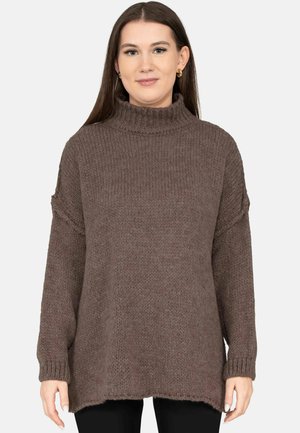 Junge Frau mit langen dunklen Haaren, die goldene Creolen und einen groben braunen Rollkragenpullover trägt, vor einem einfarbigen Hintergrund stehend.