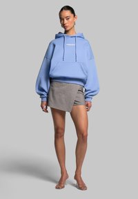 Blå hættejakke med en frontlomme og snore, parret med grå indpakningsstil shorts. Modellen bærer klare strop sandaler. Glatte teksturer.