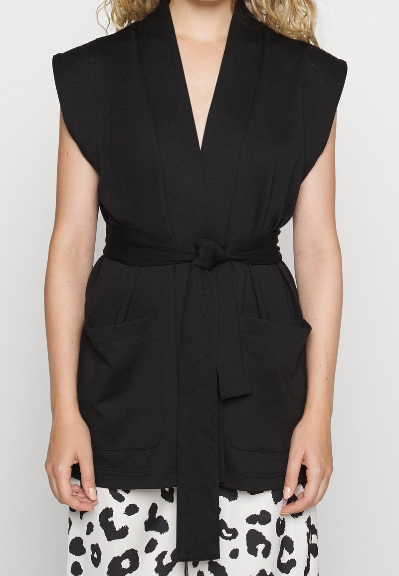 Femme portant un gilet noir sans manches ceinturé avec des poches sur un pantalon à imprimé animal noir et blanc.