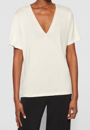 Personne portant un haut blanc à col en V, à manches courtes et ample, et un pantalon noir, se tenant avec les mains détendues le long du corps.