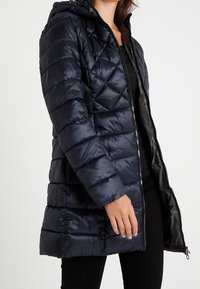 Navy blue puffy coat with a zip closure, quilted design, detachable hood, and long sleeves. Made of smooth, shiny fabric.