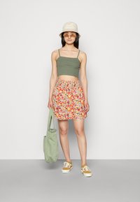 Top cropped verde, saia com padrão floral e cintura elástica, chapéu de balde bege e ténis amarelos. Modelo segura uma bolsa tote verde-claro.