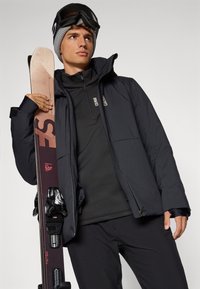 Schwarze isolierte Jacke mit hohem Kragen, kombiniert mit einem schwarzen Thermo-Oberteil und einer Hose. Das Model hält einen braunen Ski mit Bindungen und trägt eine graue Mütze sowie eine Skibrille.