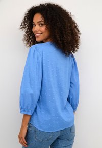 Svetlomodrá blúzka s textúrovanými vzormi v tvare srdca, nadýchanými dlhými rukávmi a voľným strihom, doplnená svetlými rifľami.