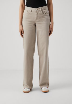 Osoba ubrana w beżowe spodnie o prostych nogawkach z przednimi kieszeniami i białe trampki, stojąca na tle jednolitego jasnego tła.