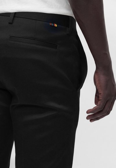 Gros plan d'une personne portant un pantalon noir avec une petite étiquette multicolore près de la poche arrière et une chemise blanche rentrée.