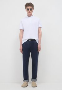 Polo shirt bianco a maniche corte con colletto alto, abbinato a pantaloni blu navy. Il modello indossa scarpe slip-on beige e occhiali da sole.