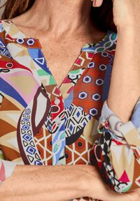 Kleurrijke, gemusterde blouse met een V-hals en knoopsluiting. De kleuren zijn rood, blauw, groen en bruin met geometrische ontwerpen.