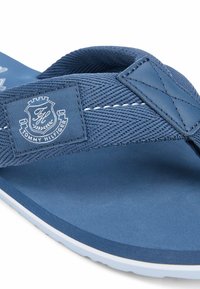 Plano detalle de una sandalia tipo chancla azul de Tommy Hilfiger con una correa de tela texturizada y un parche con el logo blanco en el lateral.