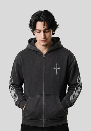 Jeune homme aux cheveux noirs ondulés portant un sweat à capuche noir zippé avec une croix sur la poitrine et des motifs de flammes sur les deux manches, les mains dans les poches.