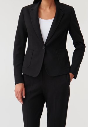 Kvinde iført en tætsiddende sort blazer over en hvid top, kombineret med matchende sorte bukser, en hånd i lommen, mod en ensfarvet baggrund.