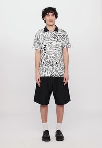 Camisa polo de meia manga em branco com design de texto abstrato em preto, combinada com calções pretos largos. O modelo está de pé sobre um fundo neutro.