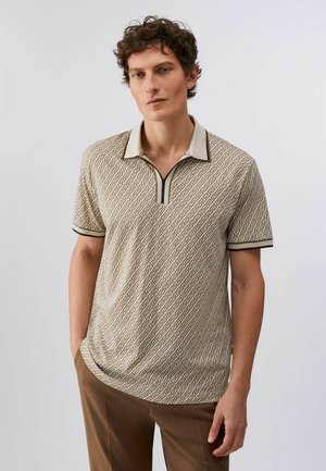 Uomo con capelli ricci castani che indossa una polo beige con motivi e bordi scuri e pantaloni marroni, in piedi contro uno sfondo chiaro e semplice.