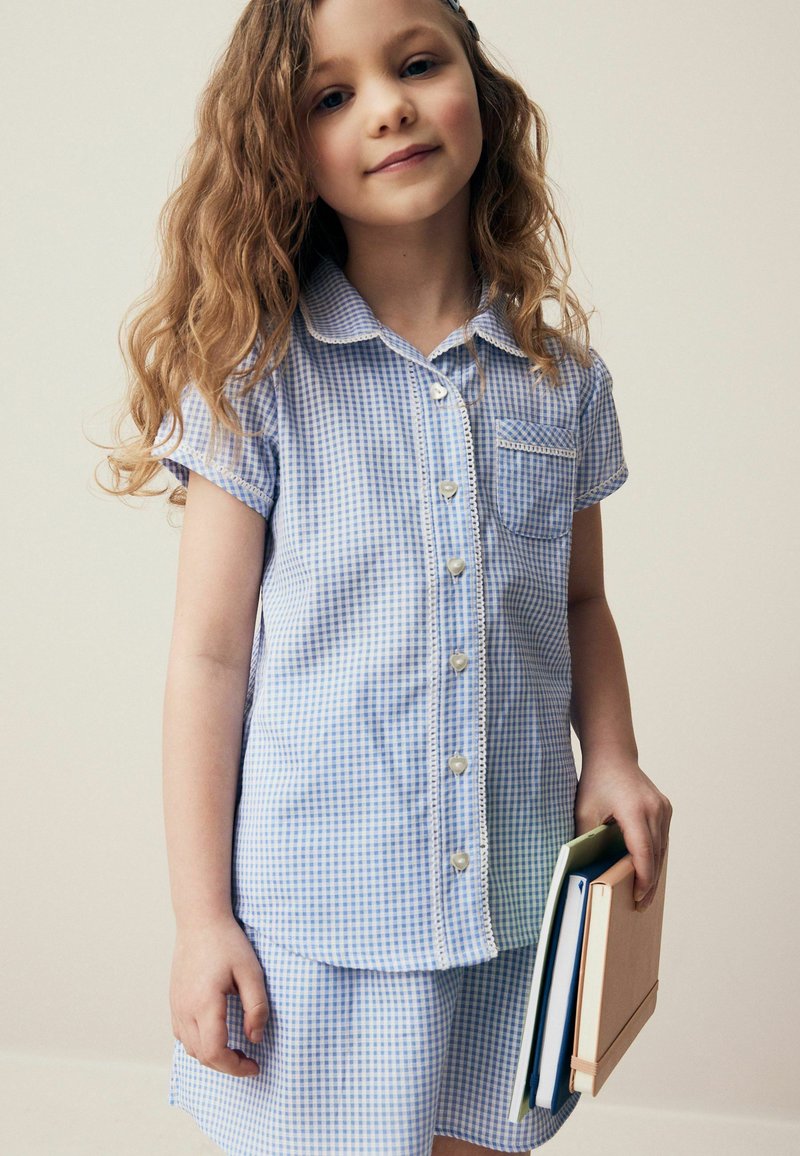 Blauw-wit geruite blouse met korte mouwen, knoopsluiting, kraag en zak. Gecombineerd met een bijpassende rok; boeken in de hand.