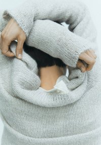 Personne tirant un pull en tricot gris clair par-dessus la tête, les mains tenant le tissu, avec des cheveux courts et foncés et un t-shirt blanc visible en dessous.