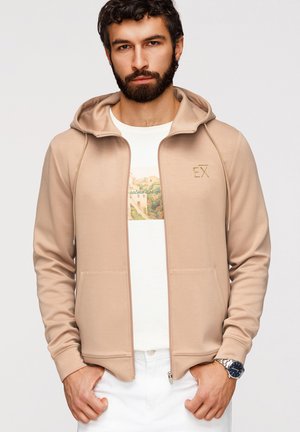 Hombre con barba que lleva una sudadera beige con cremallera, camiseta blanca con diseño gráfico, pantalones blancos y reloj de pulsera plateado, de pie con las manos en los bolsillos.