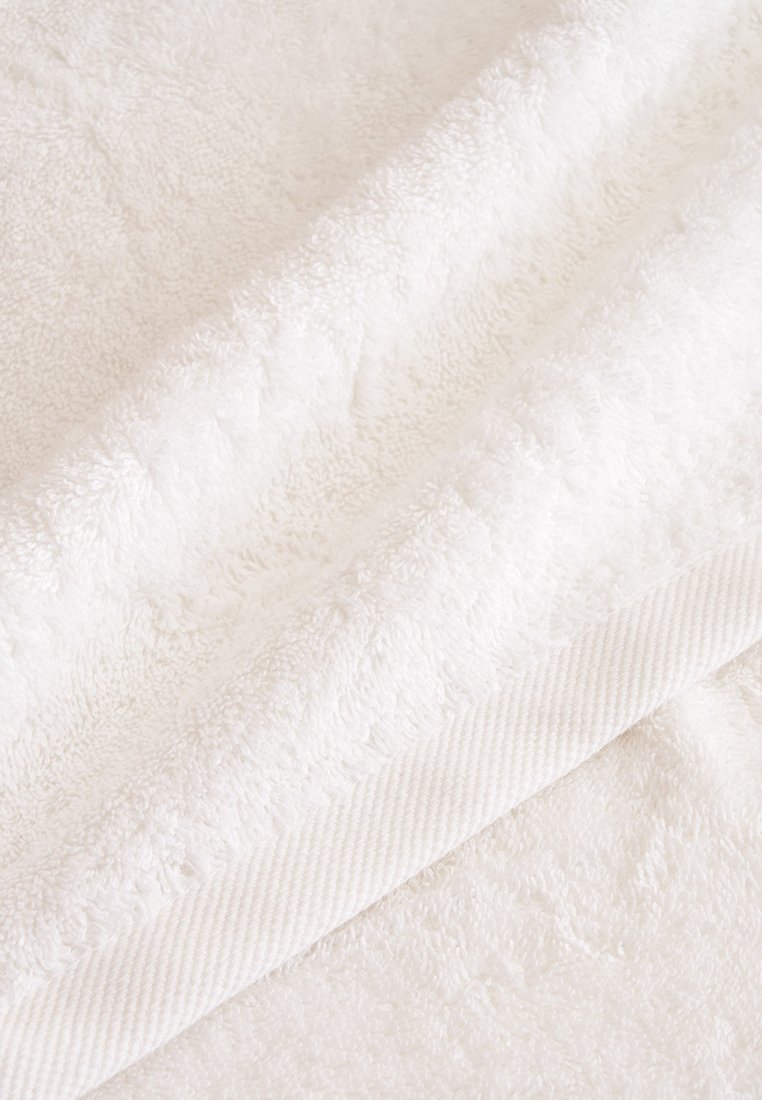 Asciugamano bianco in cotone con una superficie pelosa e strutturata e un bordo decorato con una trama tessuta, che mostra un design pulito e minimalista.
