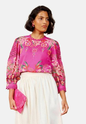 Frau trägt eine leuchtend pinke Blumenbluse mit Spitzenärmeln und einen weißen Rock, hält in ihrer linken Hand eine strukturierte pinke Clutch.