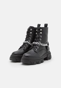 Svarta läderankelboots med en kraftig sula, prydda med silverfärgade kedjedetaljer och metallöglor, texturerade snören och en bakre dragflik.