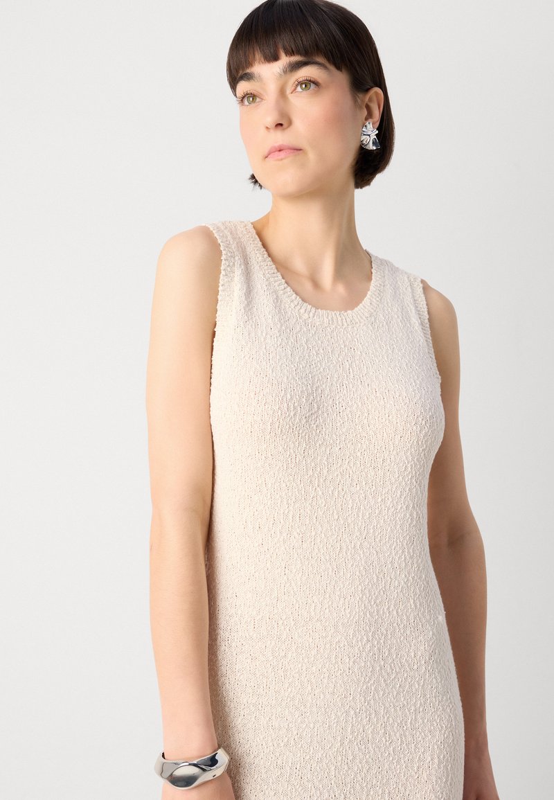 Robe sans manches beige en tissu texturé, décolleté rond et coupe slim. Présente un bracelet en argent au poignet.