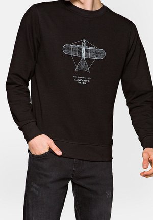 Uomo con felpa nera con disegno di un aereo vintage e testo "Taïzin Drachenflieger, 1879 LANÇERTO DYNAMIC" sul davanti.
