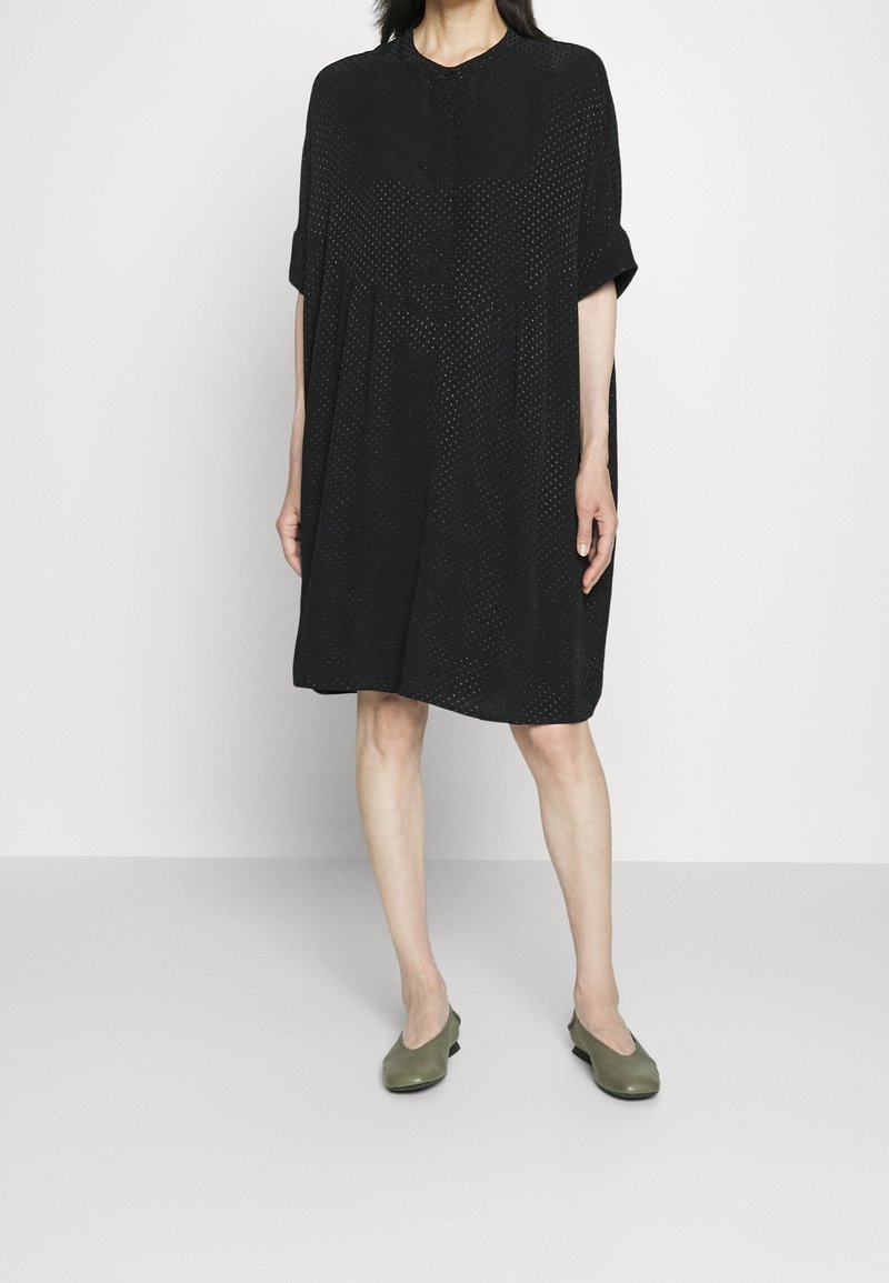 Czarna sukienka z krótkim rękawem i okrągłym dekoltem, wykonana z lekkiego, teksturowanego materiału i z plisowanym wzorem. Noszona z zielonymi balerinami.