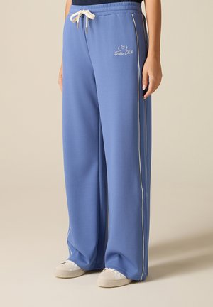 Persoon draagt losse blauwe joggingbroek met witte zijstrepen, witte trekkoord, "Relax Club" borduring en witte sneakers op een neutrale achtergrond.