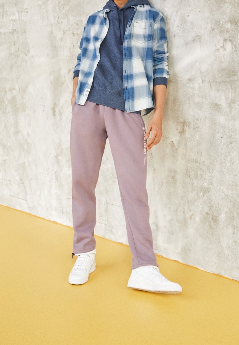 Personne portant des baskets blanches, un pantalon de survêtement mauve avec du texte sur le côté, un sweat à capuche bleu et une chemise à carreaux bleue et blanche déboutonnée.