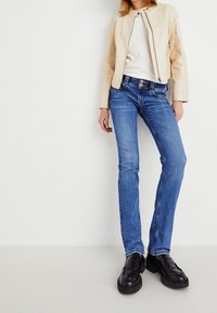 Beige läderjacka, vit skjorta, blå denimjeans med rak passform och svarta loafers. Minimal hårdvara och detaljer.