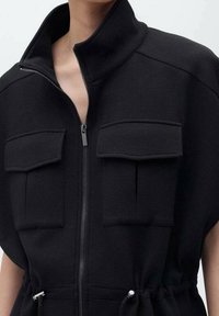 Vêtement noir à fermeture éclair doté de deux poches poitrine, d'un col montant et d'une taille réglable avec cordon. Tissu doux à texture lisse.