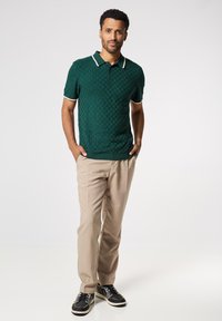 Camisa polo texturizada verde con cuello y detalles en blanco, combinada con pantalones beige y zapatos negros. Enfoque en el patrón a cuadros y el ajuste.