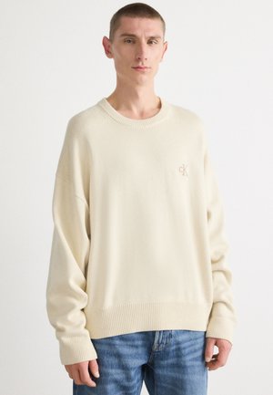 Sweater créme avec des poignets et un ourlet côtelés, arborant un logo brodé subtil, associé à un jean bleu clair. Tissu doux et texturé.