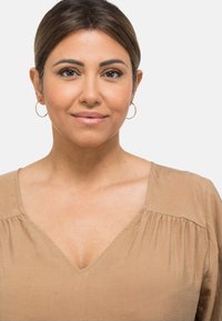 Beige V-halsbluse med rynket detalj ved skuldrene, myk tekstur og lette stoff. Modellen har på seg små hoop-øredobber.