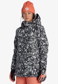 Black and white patterned snowboard jacket with a hood, featuring a logo patch. Paired with an orange beanie and black gloves.