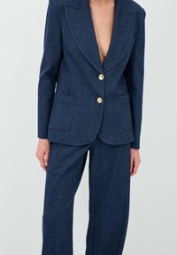 Granatowy garnitur dżinsowy z dopasowaną marynarką o głębokim dekolcie w kształcie V, złotymi guzikami oraz spodniami o szerokich nogawkach z dwiema kieszeniami.