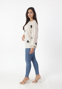 Maglione beige lavorato a maglia con motivi a stella neri, vestibilità ampia, texture a coste, abbinato a jeans skinny blu e sandali con tacco trasparente.