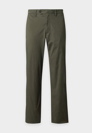 Homme debout portant une chemise sombre boutonnée avec un col beige, un pantalon vert olive et des baskets blanches sur fond blanc uni.