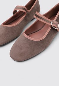 Zapatos Mary Jane planos de ante marrón con punta redondeada, acentos rosas en la correa y el forro, y un detalle de hebilla delgada. Superficie texturizada.