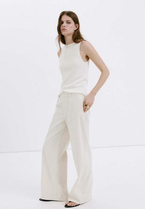 Jeune femme debout, portant un haut blanc sans manches, un pantalon crème à jambes larges, des sandales noires, avec des bagues à la main droite sur fond blanc uni.