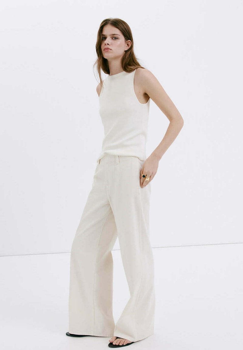 Jeune femme debout, portant un haut blanc sans manches, un pantalon crème à jambes larges, des sandales noires, avec des bagues à la main droite sur fond blanc uni.