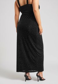 Schwarzes Maxikleid mit einem taillierten Oberteil und einem fließenden Rock im Leopardenmuster. Das Material wirkt weich mit einer subtilen Struktur. Schwarze High-Heels.