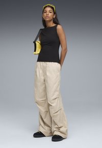 Zwarte mouwloze top, beige oversized broek, zwarte schoenen en een kleine gele tas met een zwarte band. Model staat tegen een grijze achtergrond.