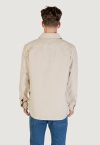 Man with curly brown hair wearing a beige long-sleeve shirt and blue jeans, shown from the back on a plain light background.