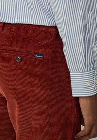 Pantalones de pana en color óxido con estrías verticales, que cuentan con un botón y una etiqueta de detalles. Combinados con una camisa de manga larga a rayas en azul claro.
