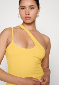 Top amarelo, ajustado, de um ombro, feito de um tecido suave. Apresenta um decote largo e tiras finas, exibindo uma silhueta elegante.