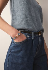 Gold bracelet featuring three circular blue stones, worn on a wrist, paired with dark denim jeans and a blue knit top with a round neckline.