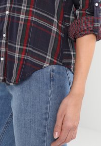 Gestreepte blouse in marineblauw, groen en rood met een zachte textuur; heeft een knoopsluiting aan de voorkant en opgerolde mouwen. Gepaard met blauwe denim jeans.