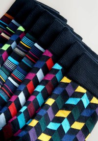 Seitse paari tumelillaid sokke, millel on värvilised geomeetrilised ja triibulised mustrid, asetatud kõrvuti heledal taustal.