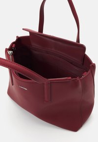 Burgundy nahkakäsilaukku, jossa on kaksi kahvaa, sisällä vetoketjullinen tasku ja useita lokeroita. Omistaa sileän pinnan ja brändin logon.